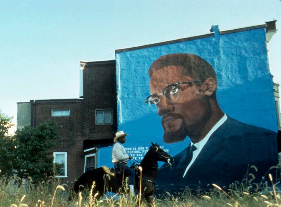 A man rides a horse in front of a mural of Malcolm X.