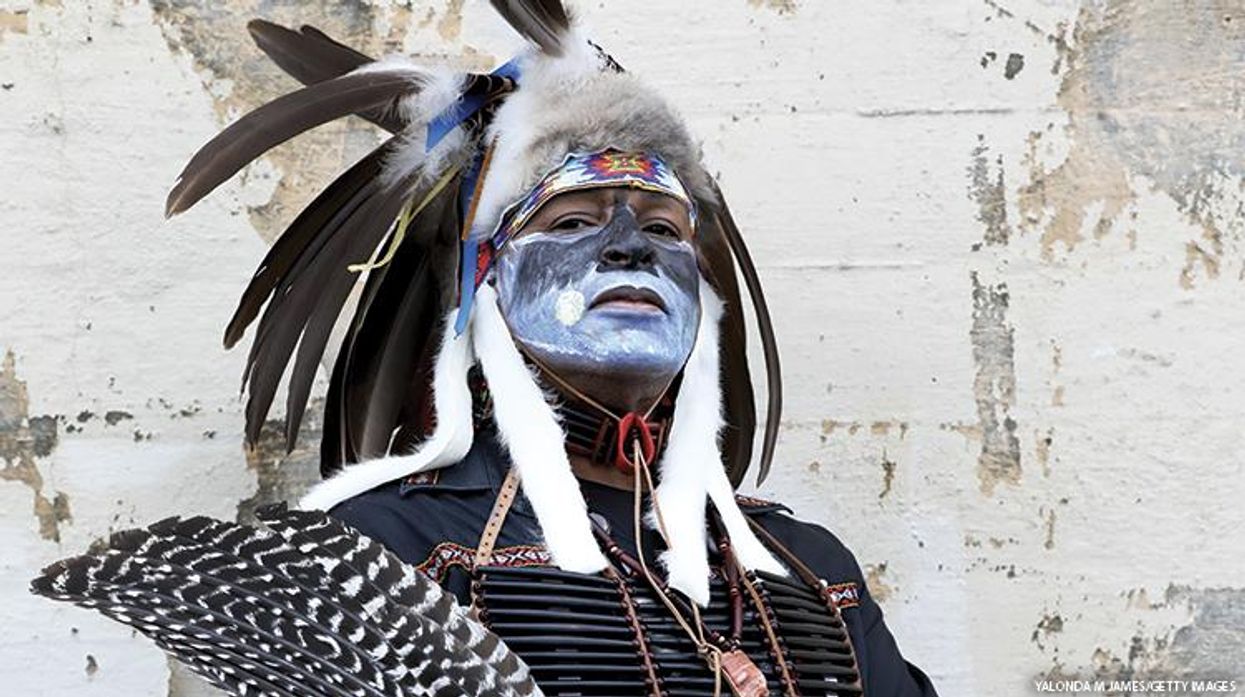 An Indigenous man in traditional garb gazes down toward the camera.