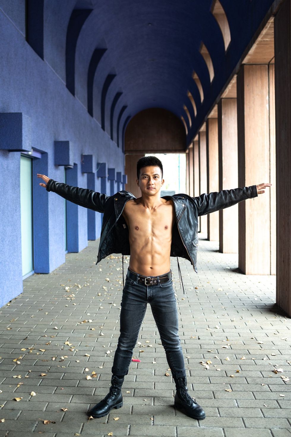 Asian American man wears jeans and a leather jacket over bare chest, stands arms outstretched