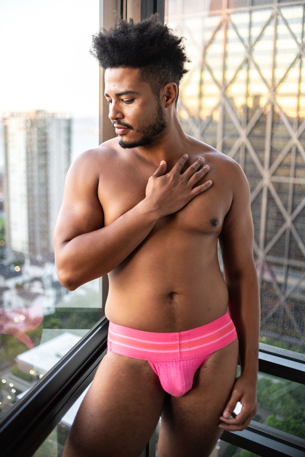 Black man stands in a jock strap with his hand on his chest in a glass elevator