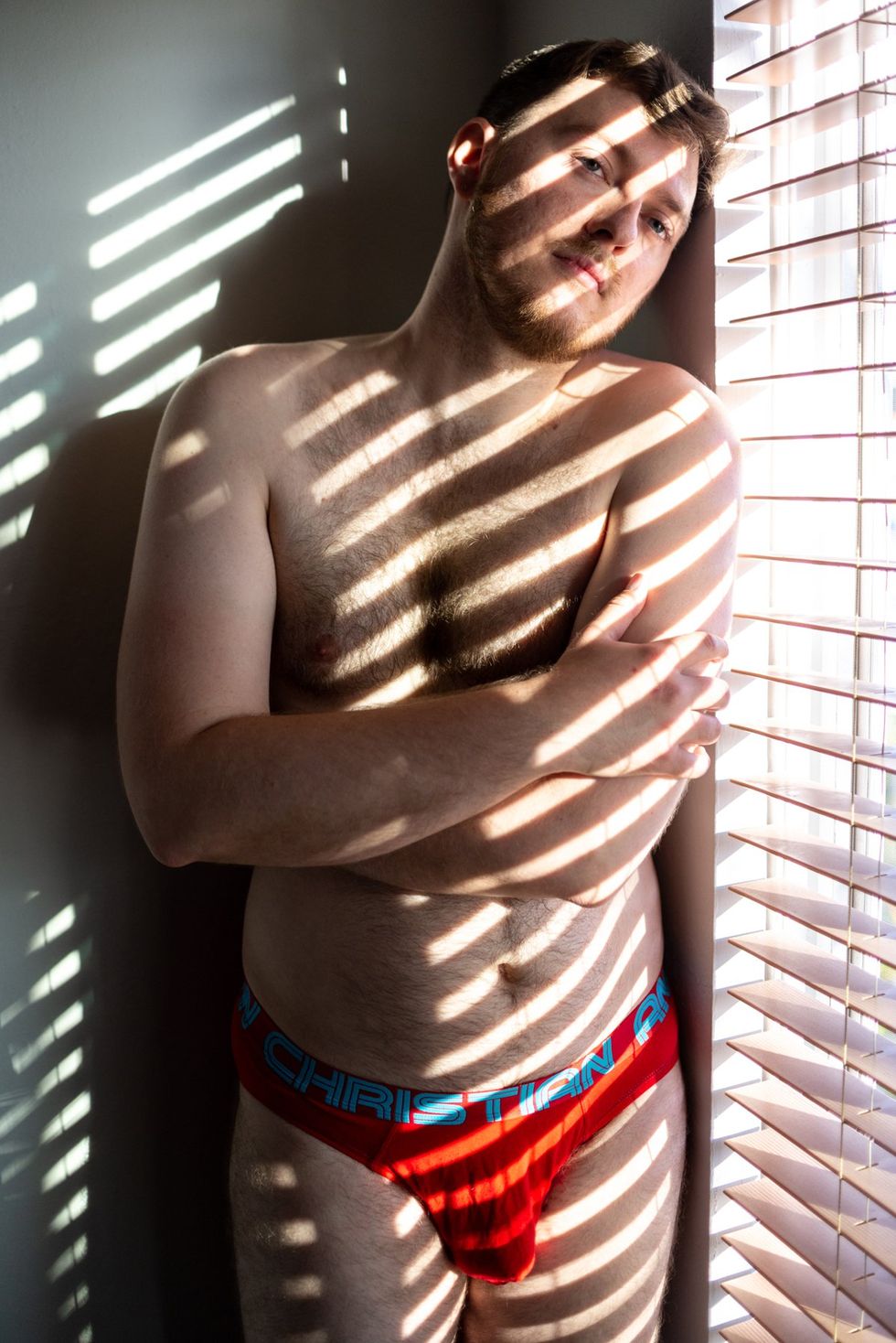 blue-eyed man with arms crossed in underwear stands by a window, shaded by blinds