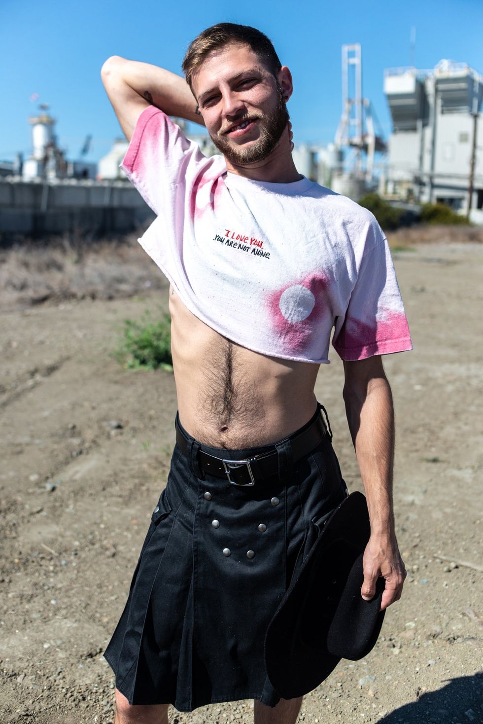 gay man in cut off t-shirt and black kilt stands outside in industrial setting