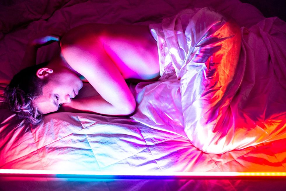 man lies shirtless in bed, lit by rainbow colors