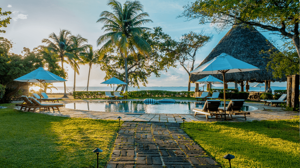 Poolside view at Turtle Inn, featuring lounge chairs, white umbrellas, palm trees, and a thatched-roof cabana overlooking the Caribbean Sea at sunset.