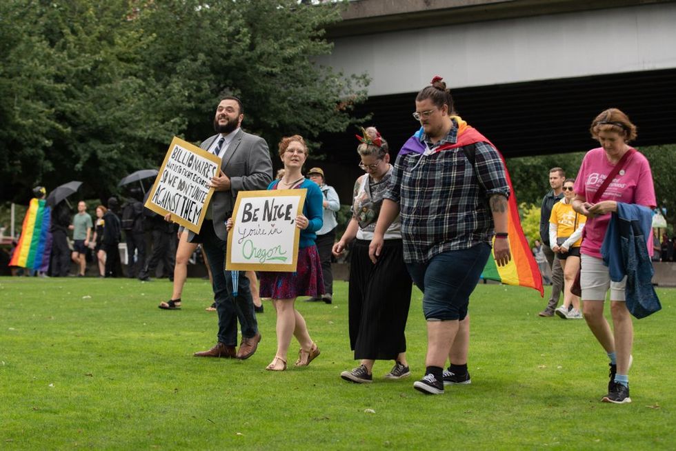 Protestors wear rainbow flag capes and carry a sign reading