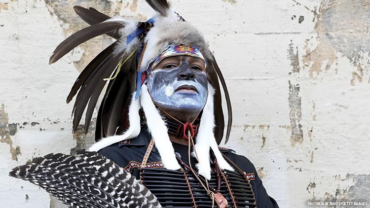 An Indigenous man in traditional garb gazes down toward the camera.